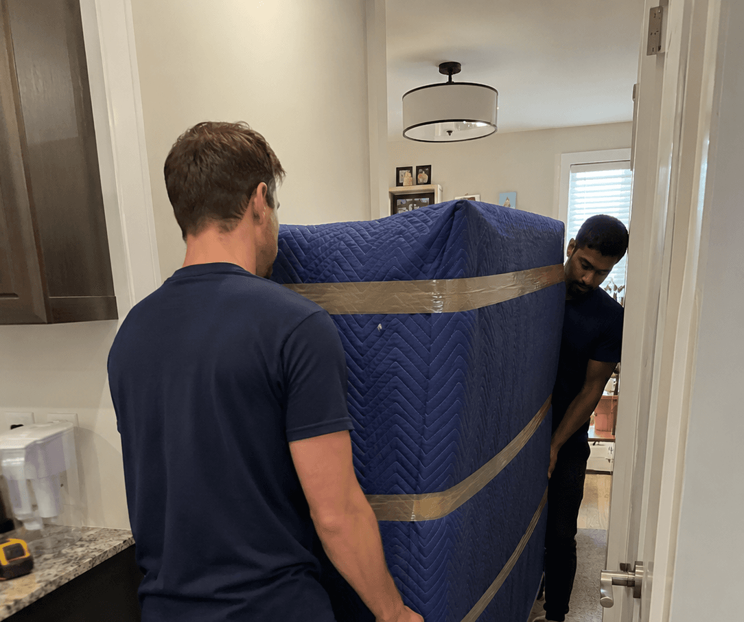Movemiles movers carrying a blanket-wrapped piece of furniture through a doorway